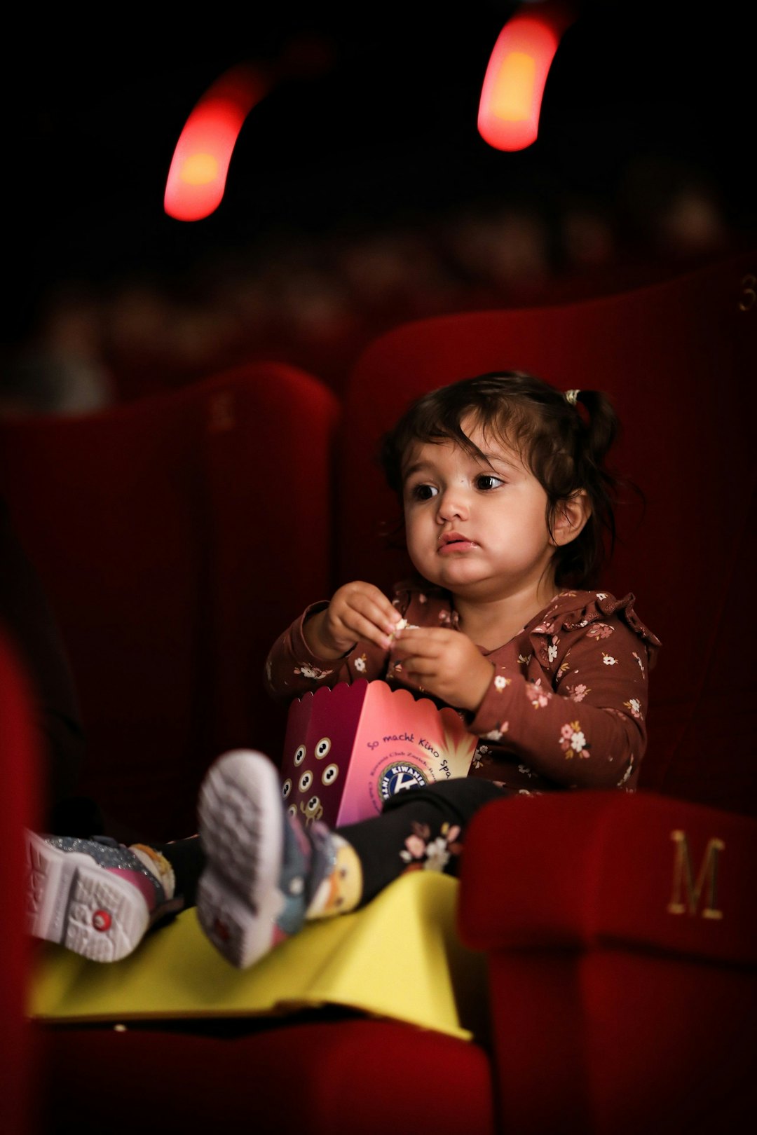 A little girl on a cinema seat eating popcorn.